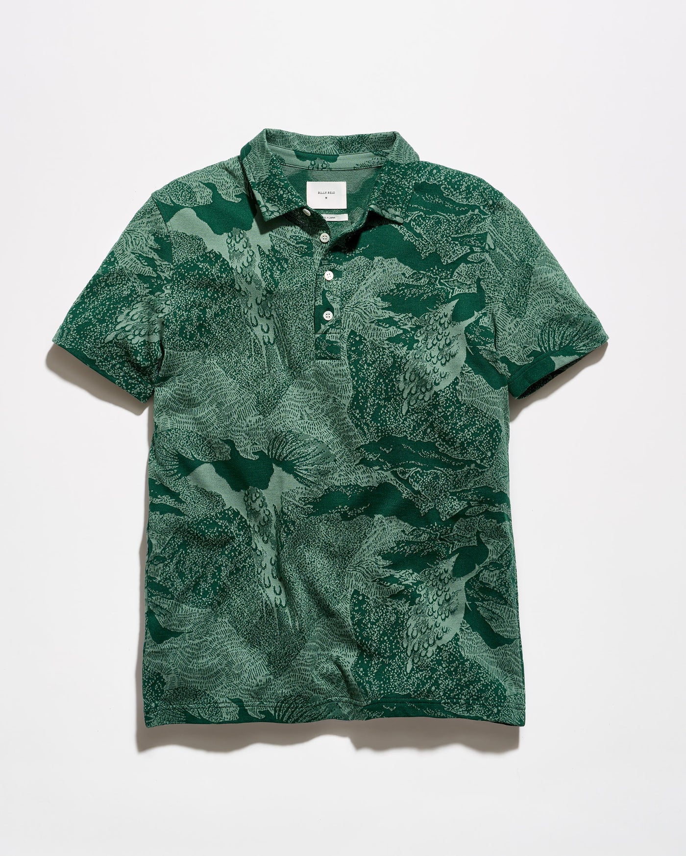 A short-sleeve polo shirt with an abstract green and white pattern, featuring a two-button placket and a classic collar, laid flat against a light gray background.