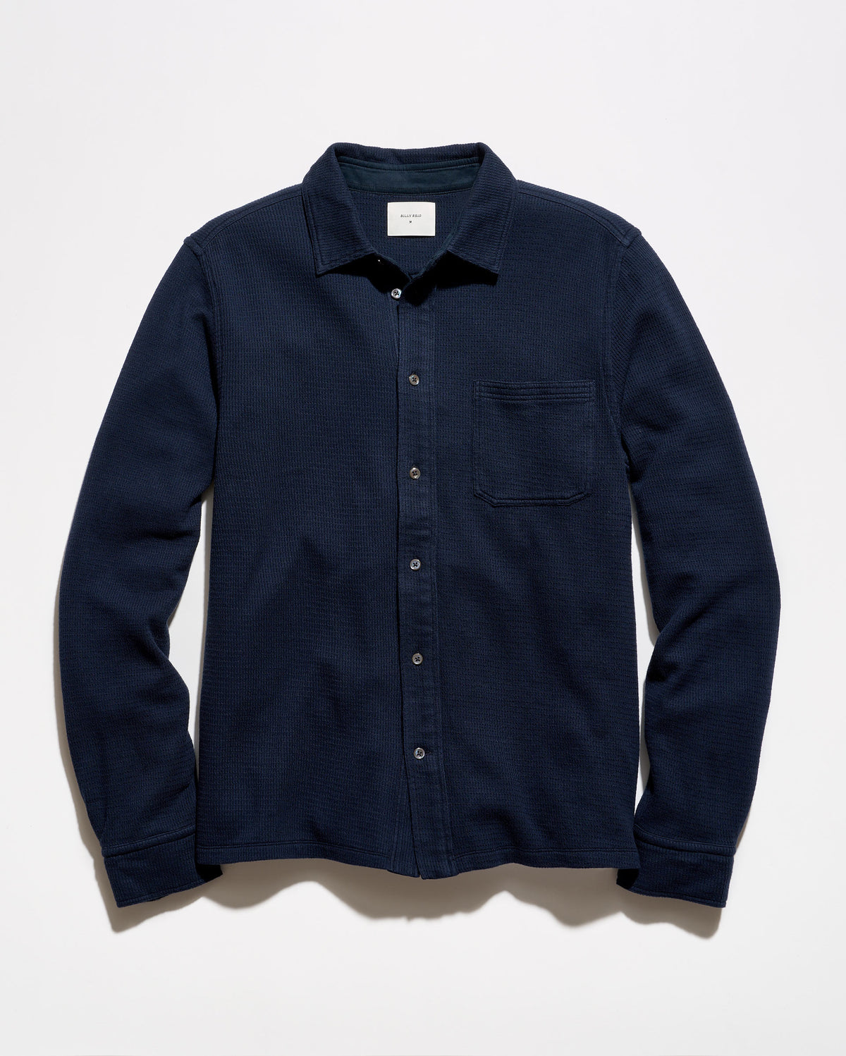 A dark navy blue long-sleeve button-up shirt with a single chest pocket, laid flat against a light off-white background.