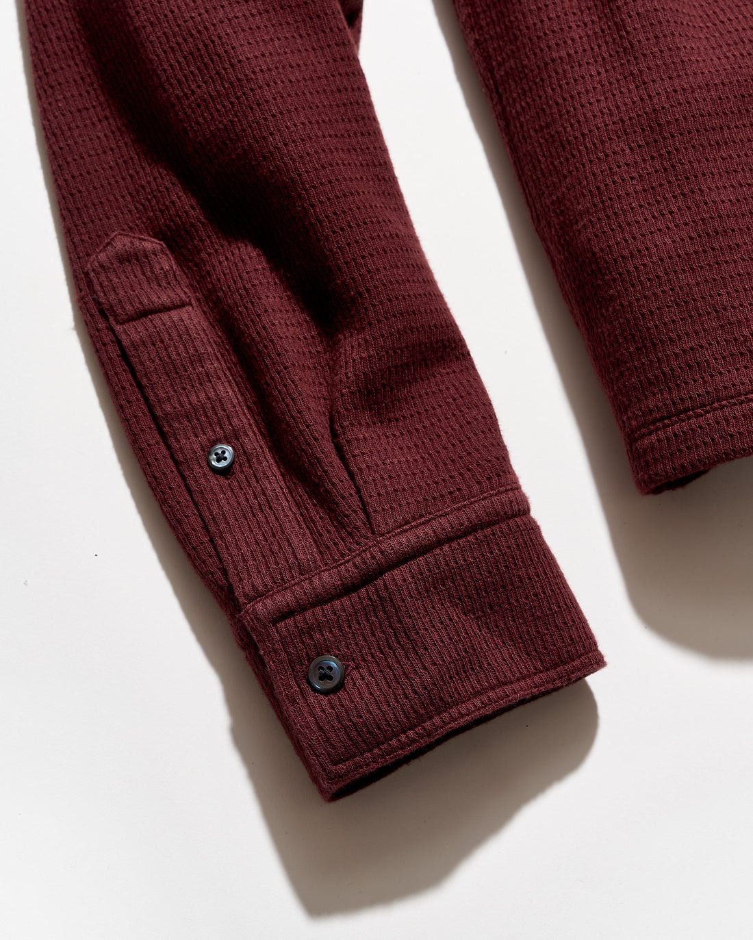 Close-up of a burgundy ladder-knit garment sleeve with a buttoned cuff, photographed on a white background.