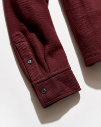 Close-up of a burgundy ladder-knit garment sleeve with a buttoned cuff, photographed on a white background.