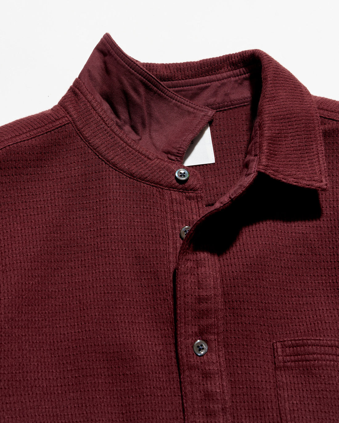 Close-up of a textured ladder-knit button down shirt in burgundy, featuring a stand-up collar, dark buttons, and a chest pocket on a white background.
