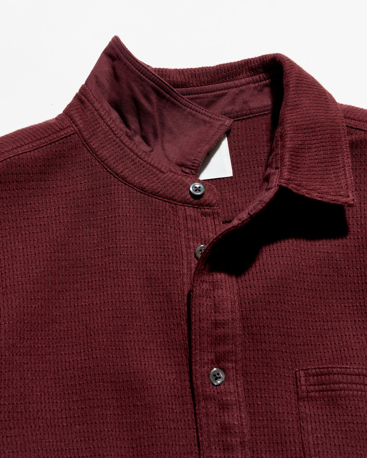 Close-up of a textured ladder-knit button down shirt in burgundy, featuring a stand-up collar, dark buttons, and a chest pocket on a white background.