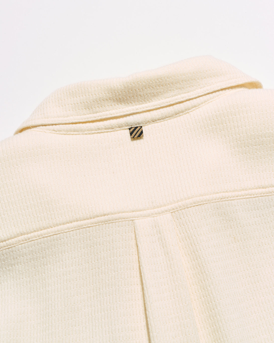 Close-up of a cream-colored textured jacket with a small gold and black striped logo tab near the collar.
