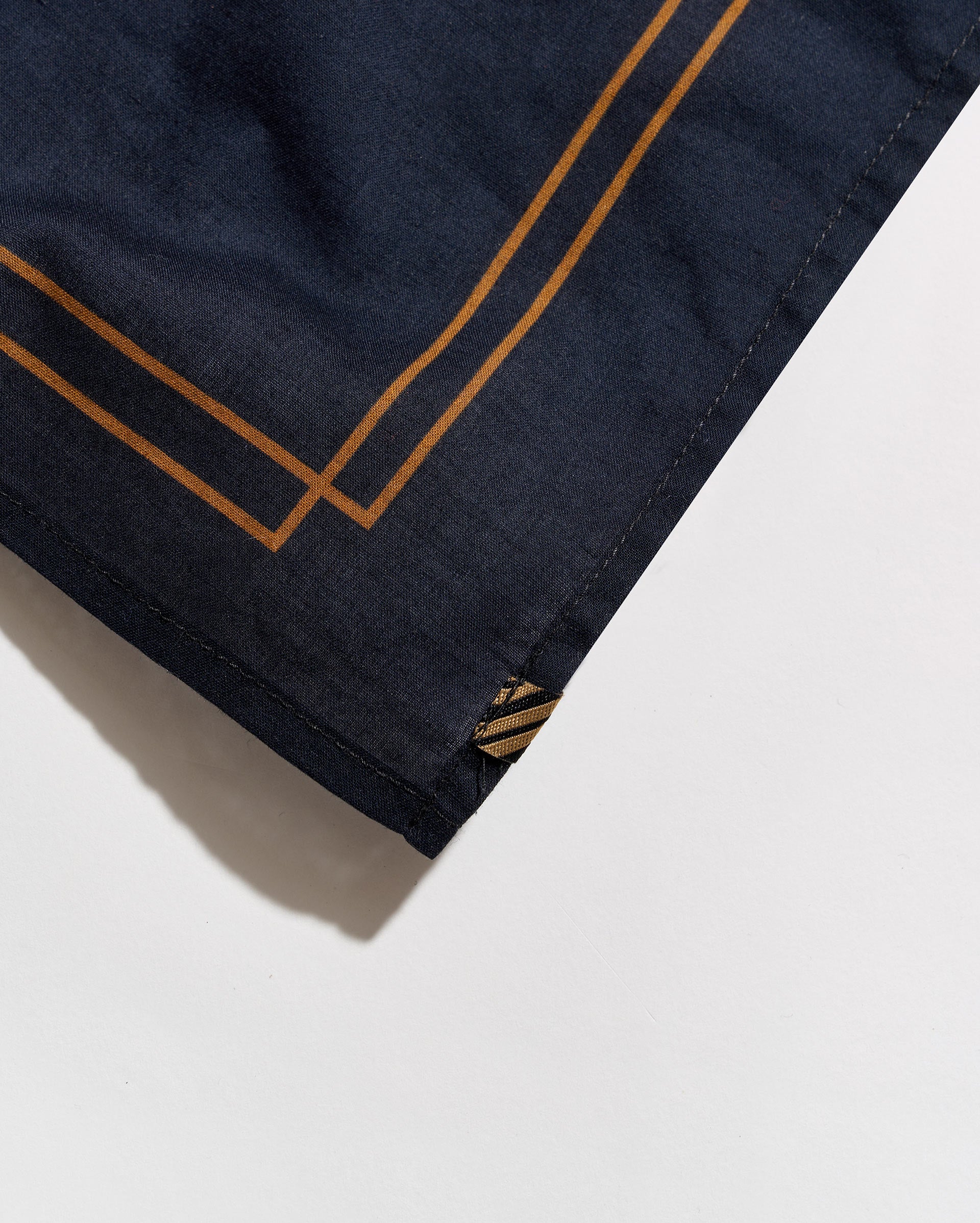 Close-up of a dark navy blue fabric product, likely a scarf or bandana, featuring double gold geometric border lines and a small gold embroidered logo at the corner, set against a clean white background.