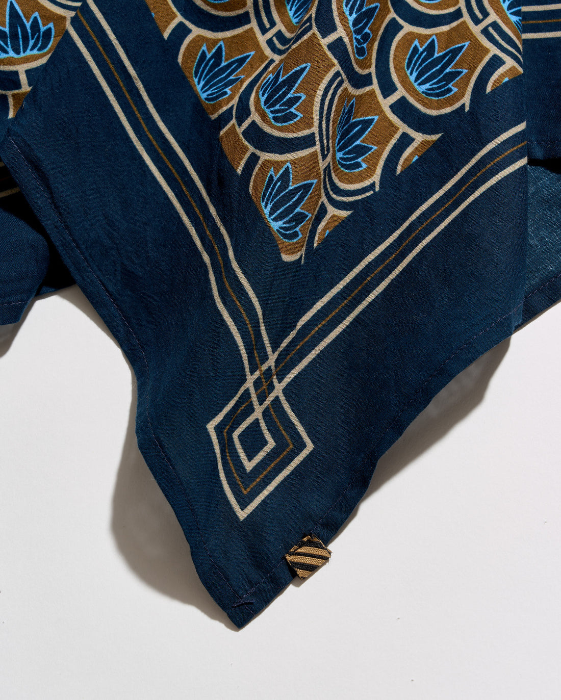 A close-up of a navy blue fabric accessory, likely a scarf or sash, featuring a detailed batik-style pattern in brown, cream, and blue. The fabric is draped diagonally on a clean white background.