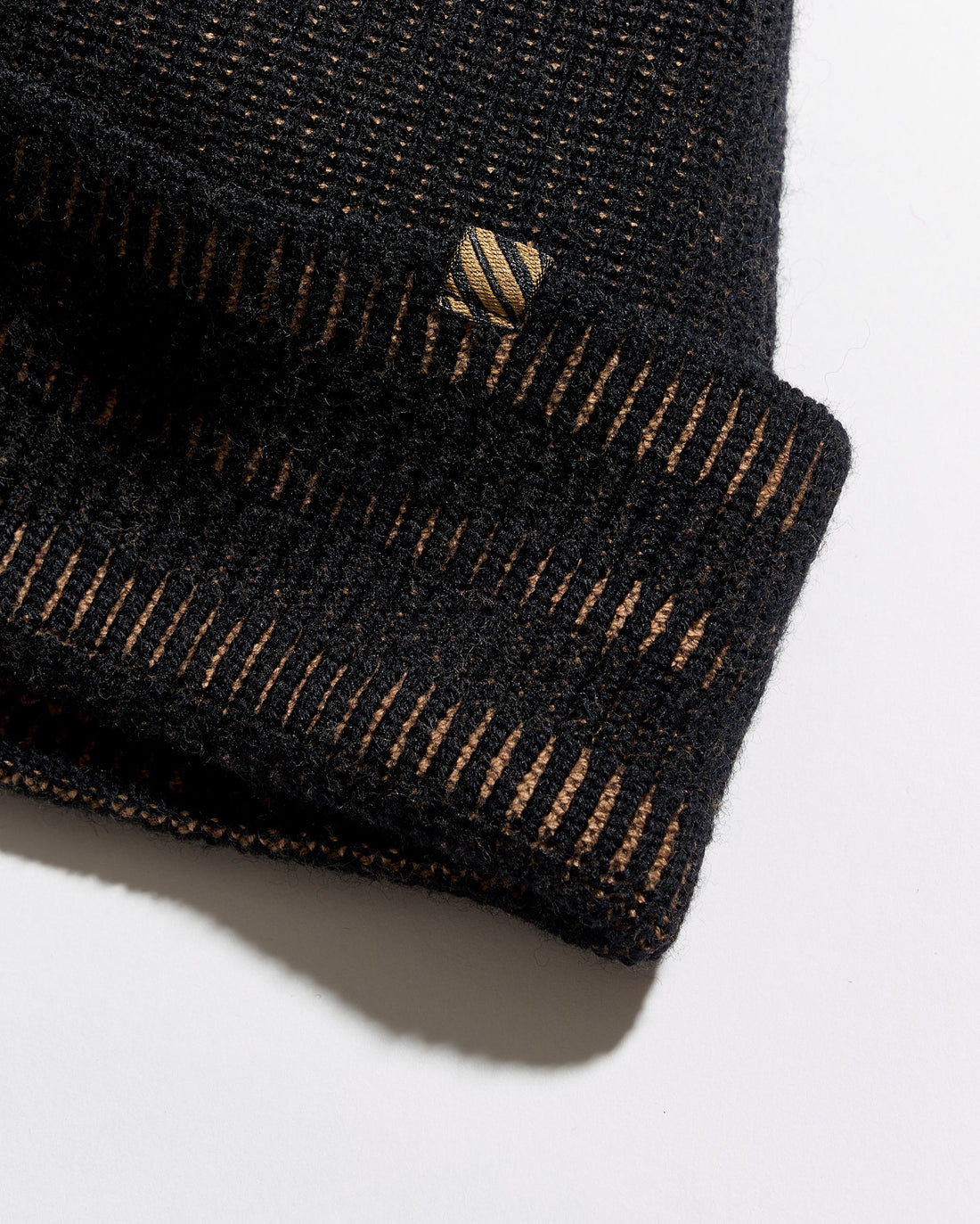 Close-up of a black knit beanie with a folded cuff, showing fine horizontal ribbing and thin gold-toned vertical stripe accents. A small embroidered logo patch is visible on the cuff.