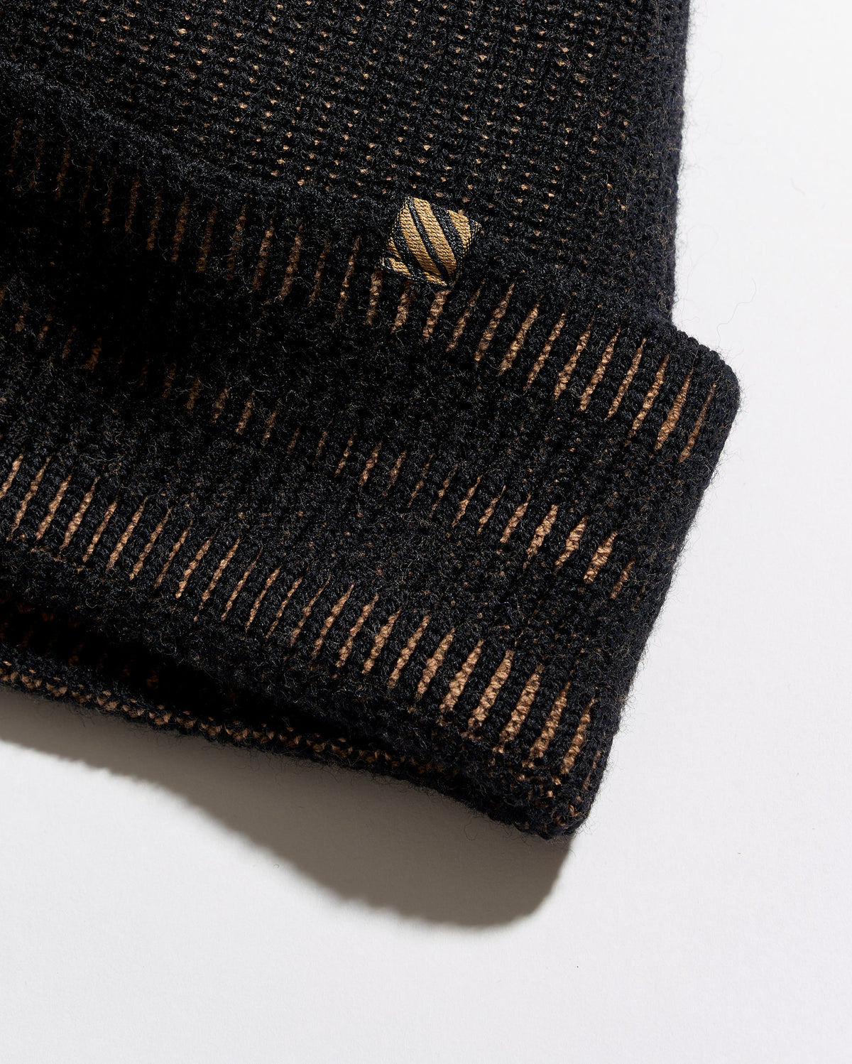 Close-up of a black knit beanie with a folded cuff, showing fine horizontal ribbing and thin gold-toned vertical stripe accents. A small embroidered logo patch is visible on the cuff.