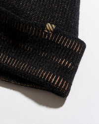 Close-up of a black knit beanie with a folded cuff, showing fine horizontal ribbing and thin gold-toned vertical stripe accents. A small embroidered logo patch is visible on the cuff.