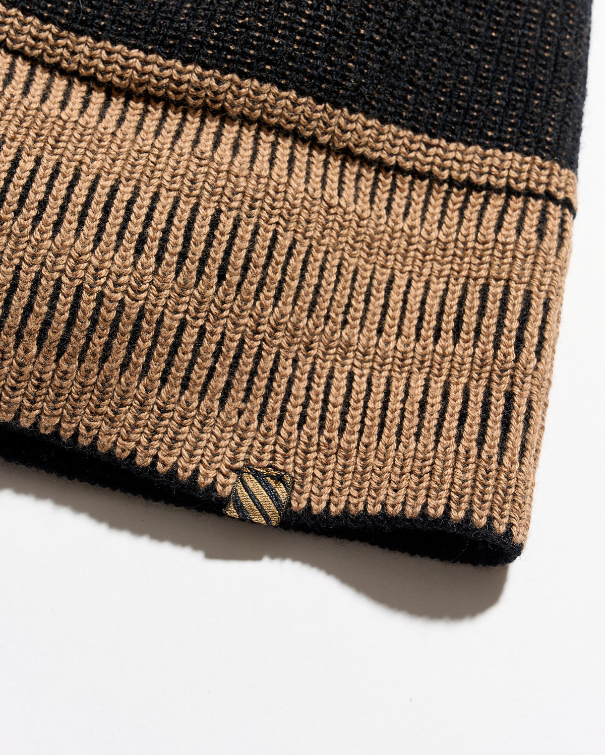 Close-up of a knitted garment cuff in black and tan with a striped accent tag near the hem, photographed on a white background.