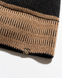 Close-up of a knitted garment cuff in black and tan with a striped accent tag near the hem, photographed on a white background.