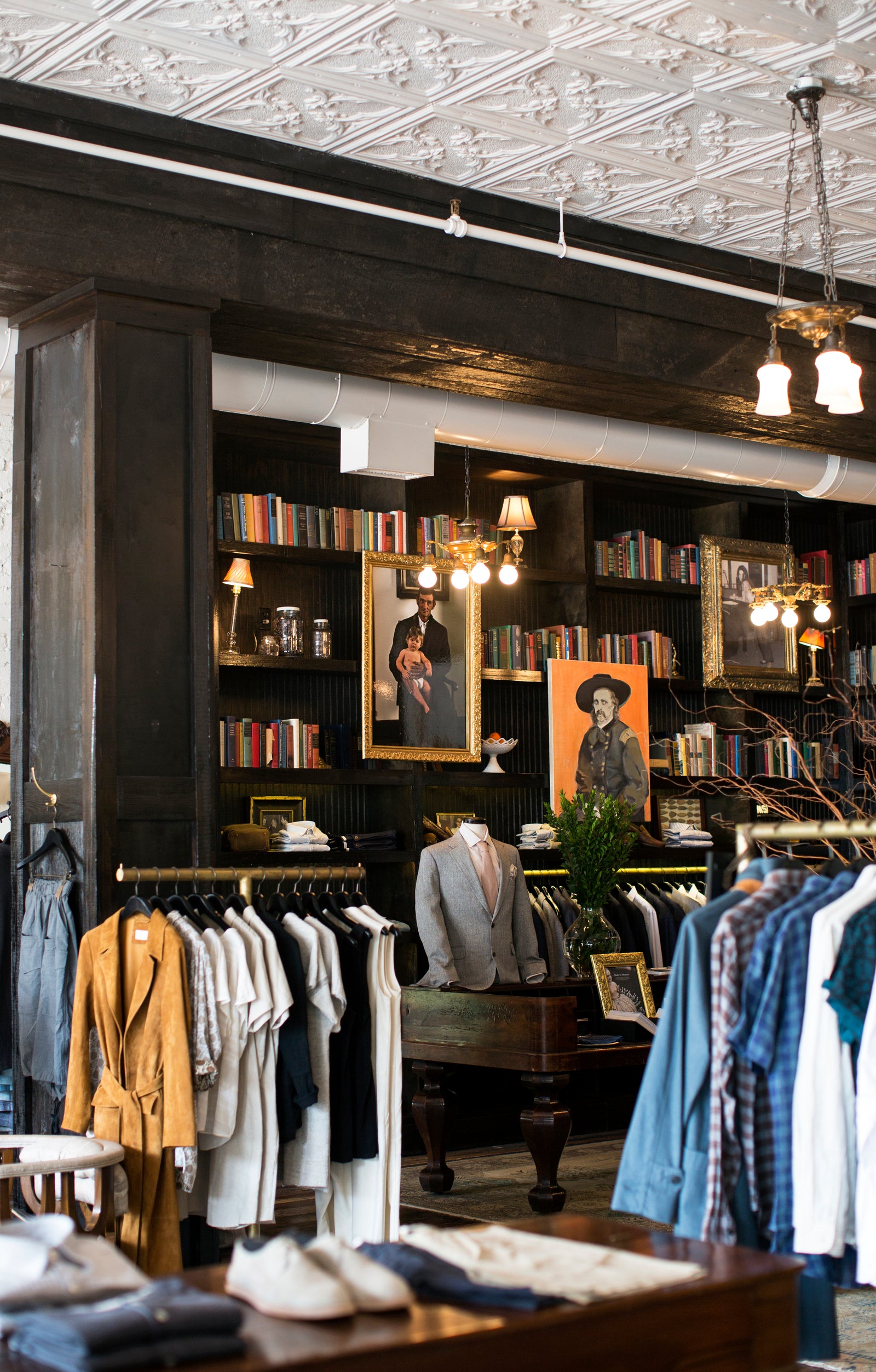 A boutique interior with clothing racks, bookshelves filled with books, framed portraits, and warm hanging lights in a dark wood and tin-ceilinged shop.