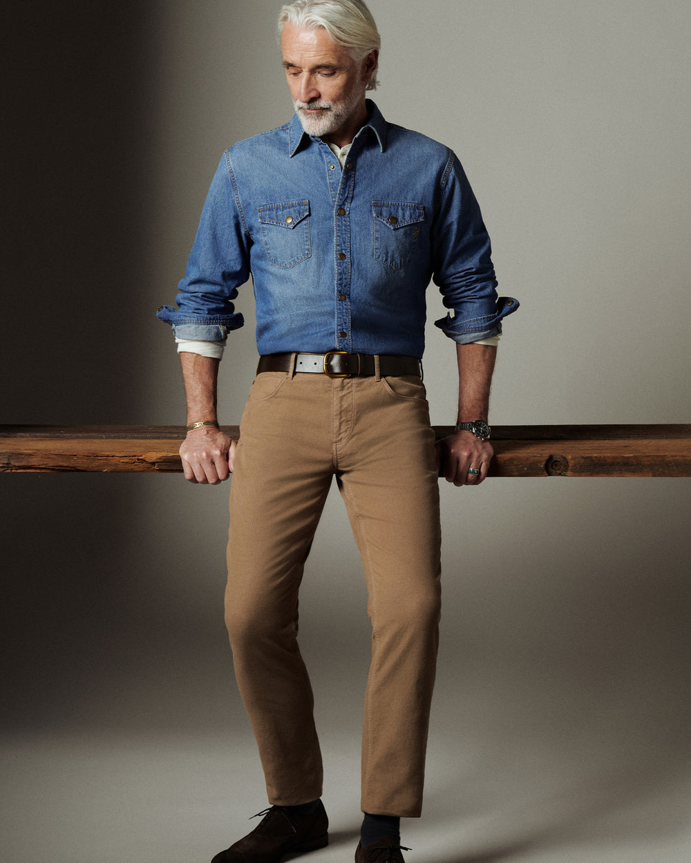 Man wearing a blue denim shirt and beige pants holding a wooden board against a neutral background