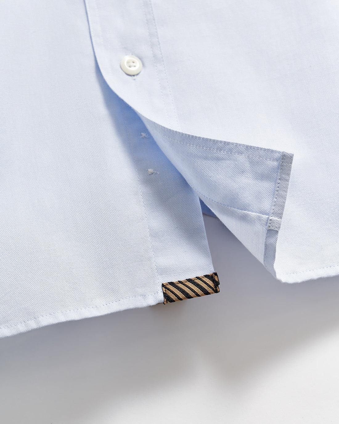 Close-up of the hem of a light blue shirt, showing a white button, a side slit, and a small black and gold striped fabric tag sewn into the bottom edge.