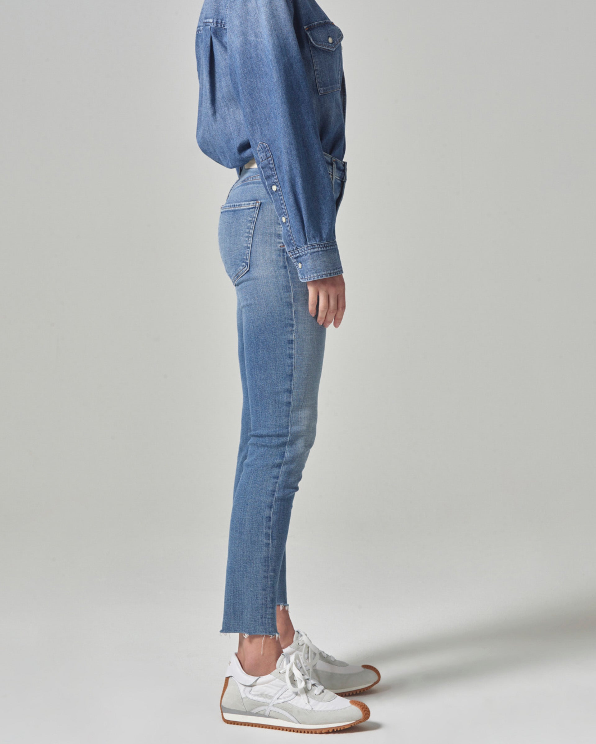 A person stands sideways wearing a denim shirt, light blue skinny jeans with frayed hems, and white sneakers with brown soles, against a plain light background.