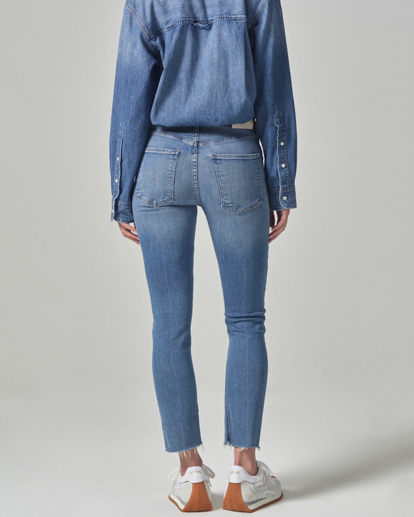 A person stands facing away, wearing a denim shirt tucked into light blue skinny jeans with raw hem and white sneakers, against a plain light background.