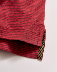 Close-up of the hem of a red, horizontally striped shirt with a small side slit. The inside hem features a contrasting black and gold diagonal striped trim. The fabric texture is clearly visible.