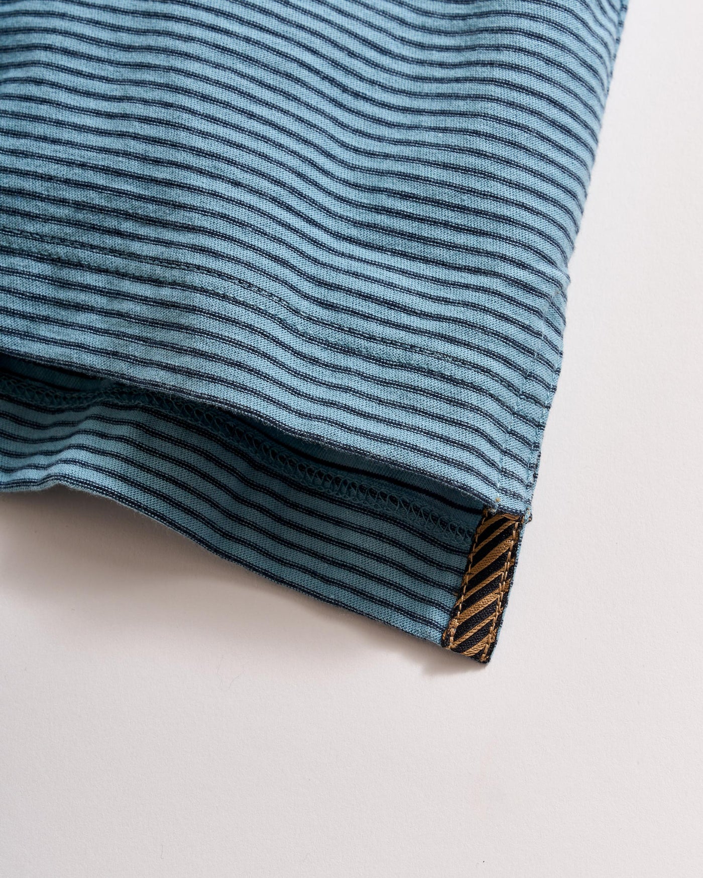 A close-up of a blue fabric with thin black horizontal stripes, showing a neatly stitched hem and a small brown tag at the edge with a diagonal pattern.