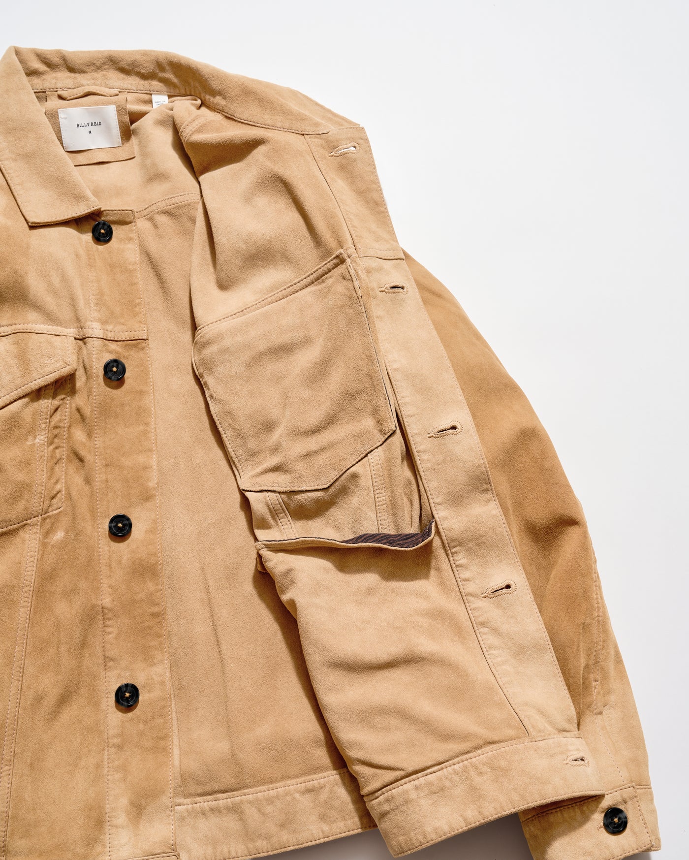 A close-up of a tan suede with black buttons, showing the collar, chest pockets, and sleeve details against a plain white background.