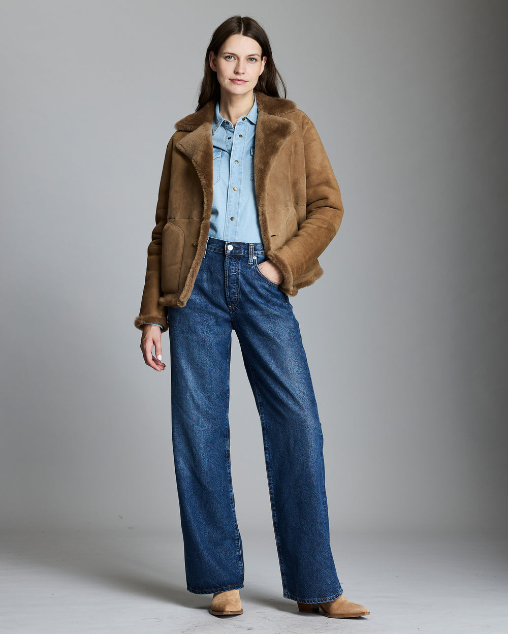 Woman wearing a brown jacket, light blue shirt, and blue jeans against a gray background