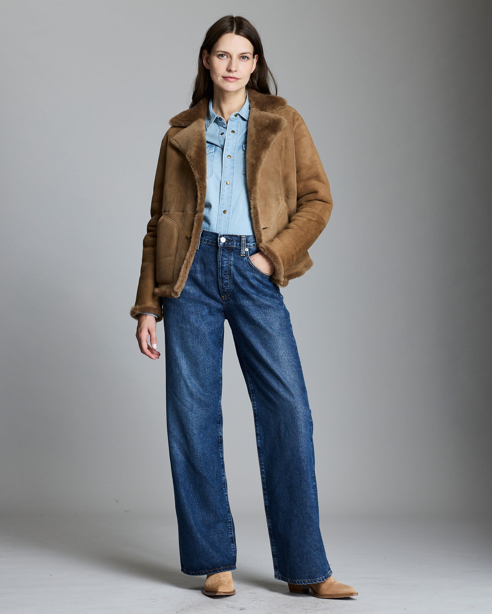 Woman wearing a brown jacket, light blue shirt, and blue jeans against a gray background