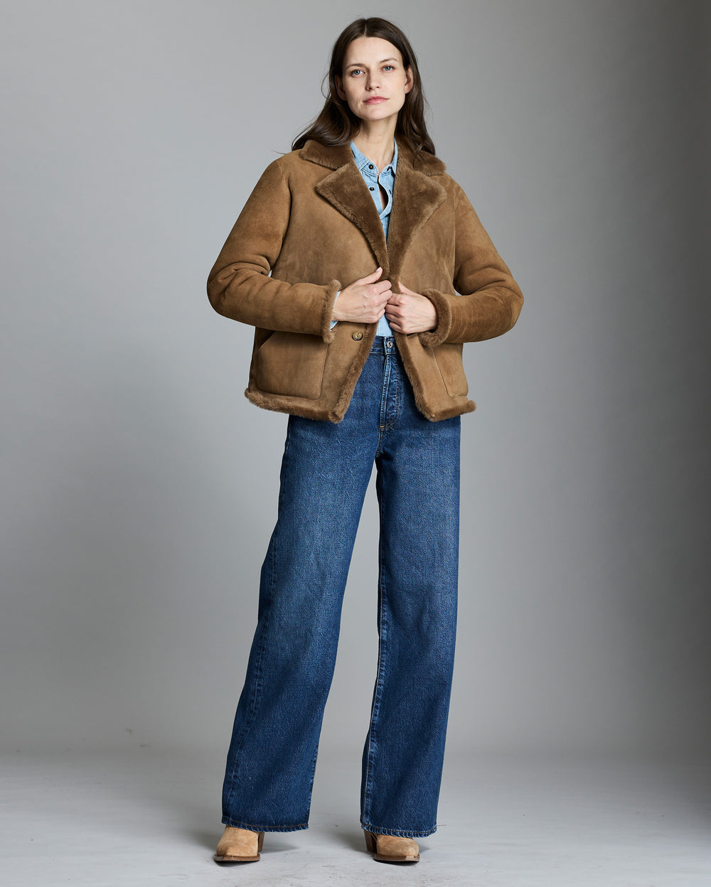 Woman wearing a brown shearling jacket and blue jeans on a gray background