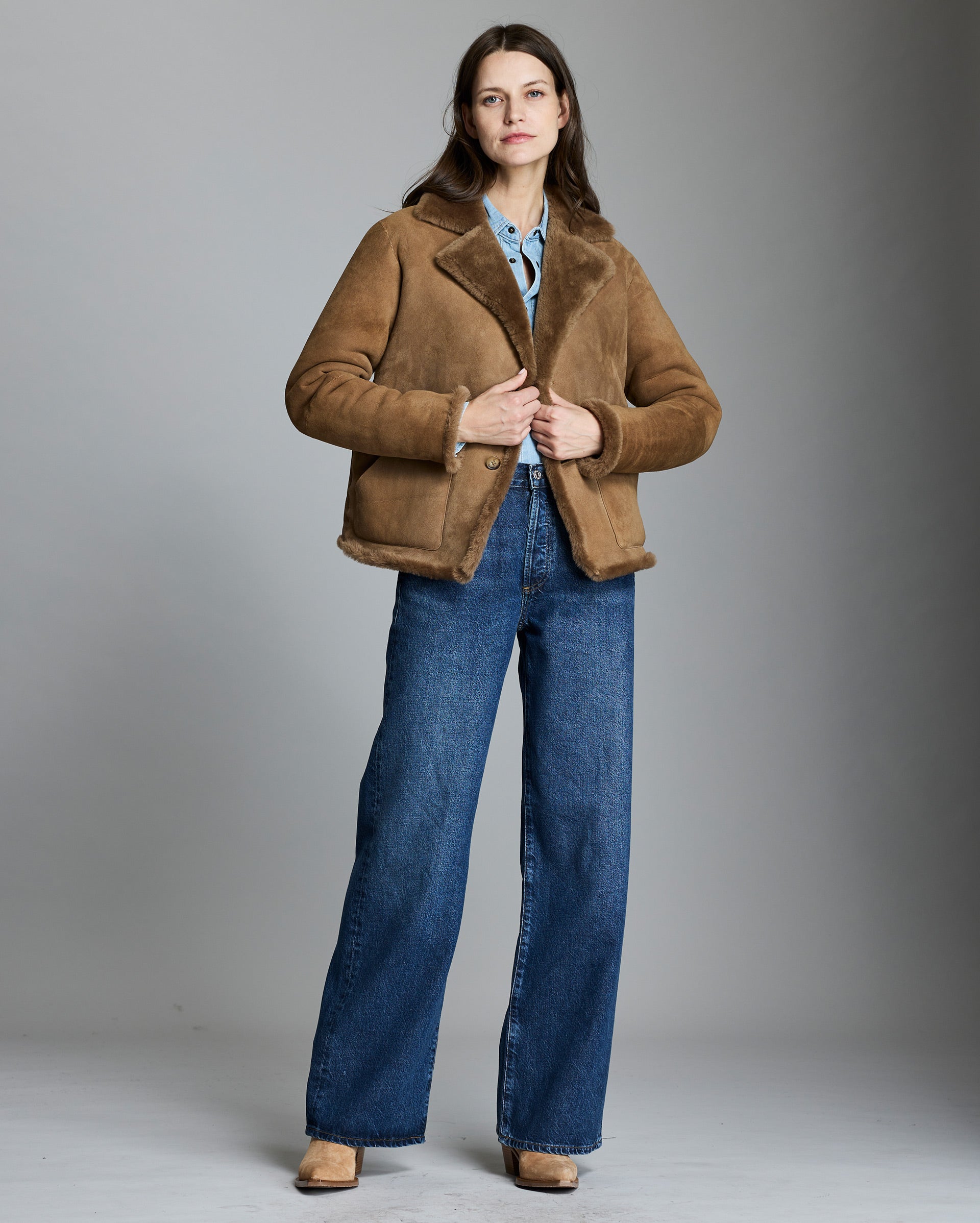 Woman wearing a brown shearling jacket and blue jeans on a gray background
