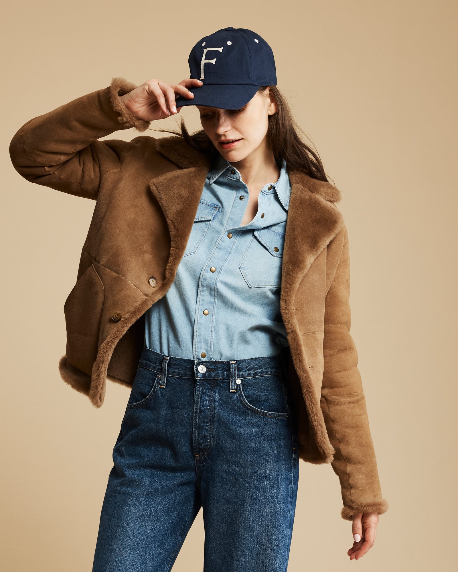 Woman wearing a brown jacket, blue denim shirt, and jeans with a navy cap on a beige background