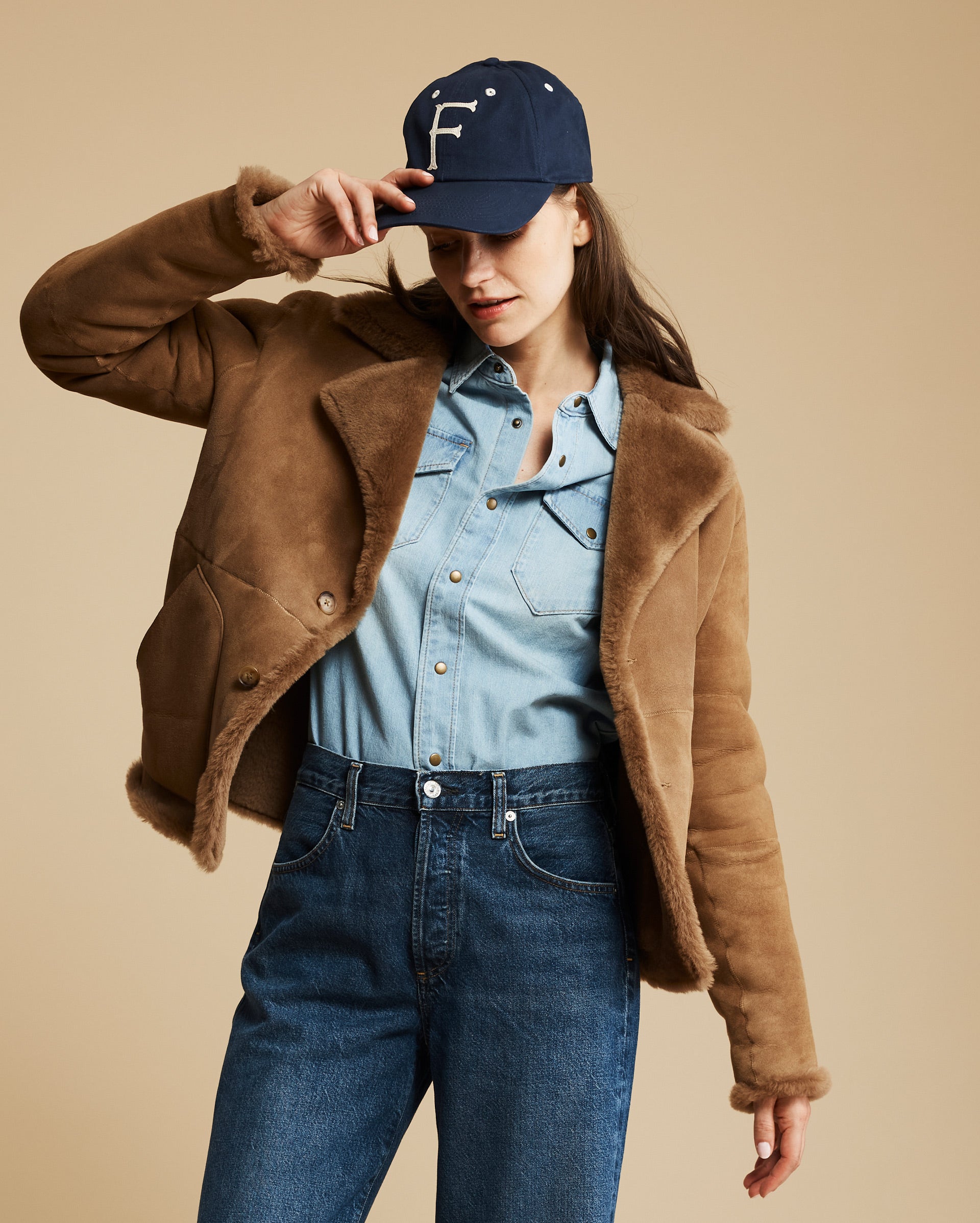 Woman wearing a brown jacket, blue denim shirt, and jeans with a navy cap on a beige background