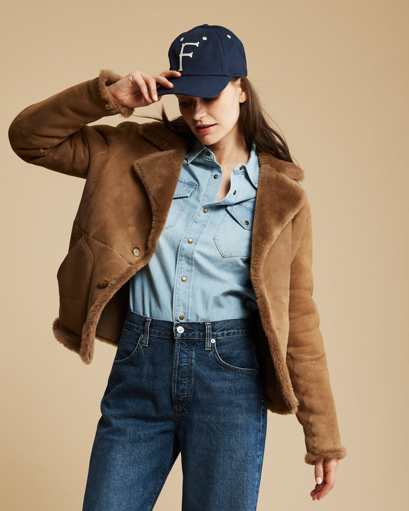 Woman wearing a brown jacket, blue denim shirt, and jeans with a navy cap on a beige background Alternate