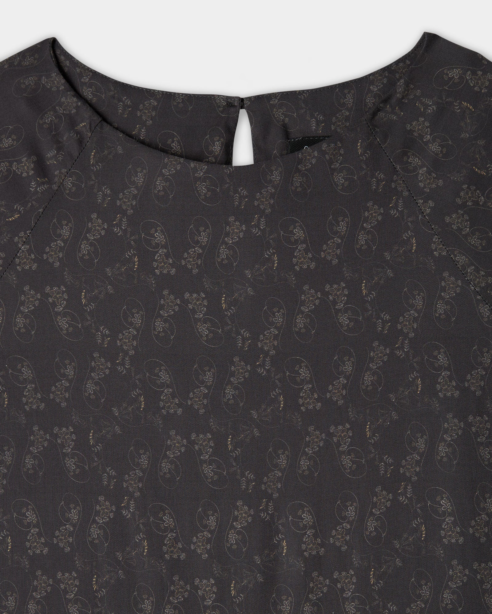 Close-up of the upper part of a dark-colored blouse with a subtle floral and paisley pattern, round neckline, and a small buttoned keyhole opening at the back.
