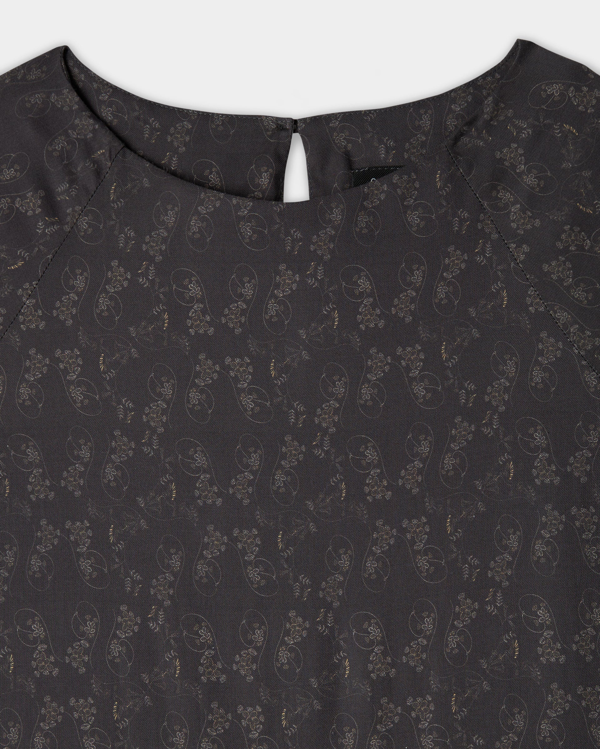 Close-up of the upper part of a dark-colored blouse with a subtle floral and paisley pattern, round neckline, and a small buttoned keyhole opening at the back.