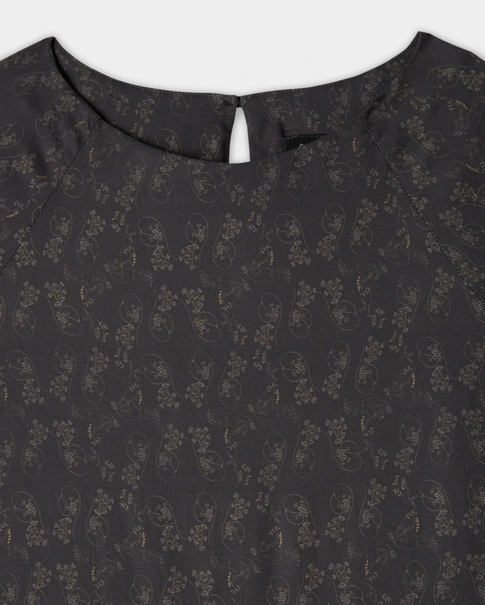 Close-up of the upper part of a dark-colored blouse with a subtle floral and paisley pattern, round neckline, and a small buttoned keyhole opening at the back.