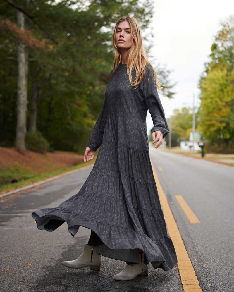 A woman with long blond hair wears a flowing, dark gray dress and light-colored boots, walking gracefully on an empty road lined with trees on a cloudy day. Alternate