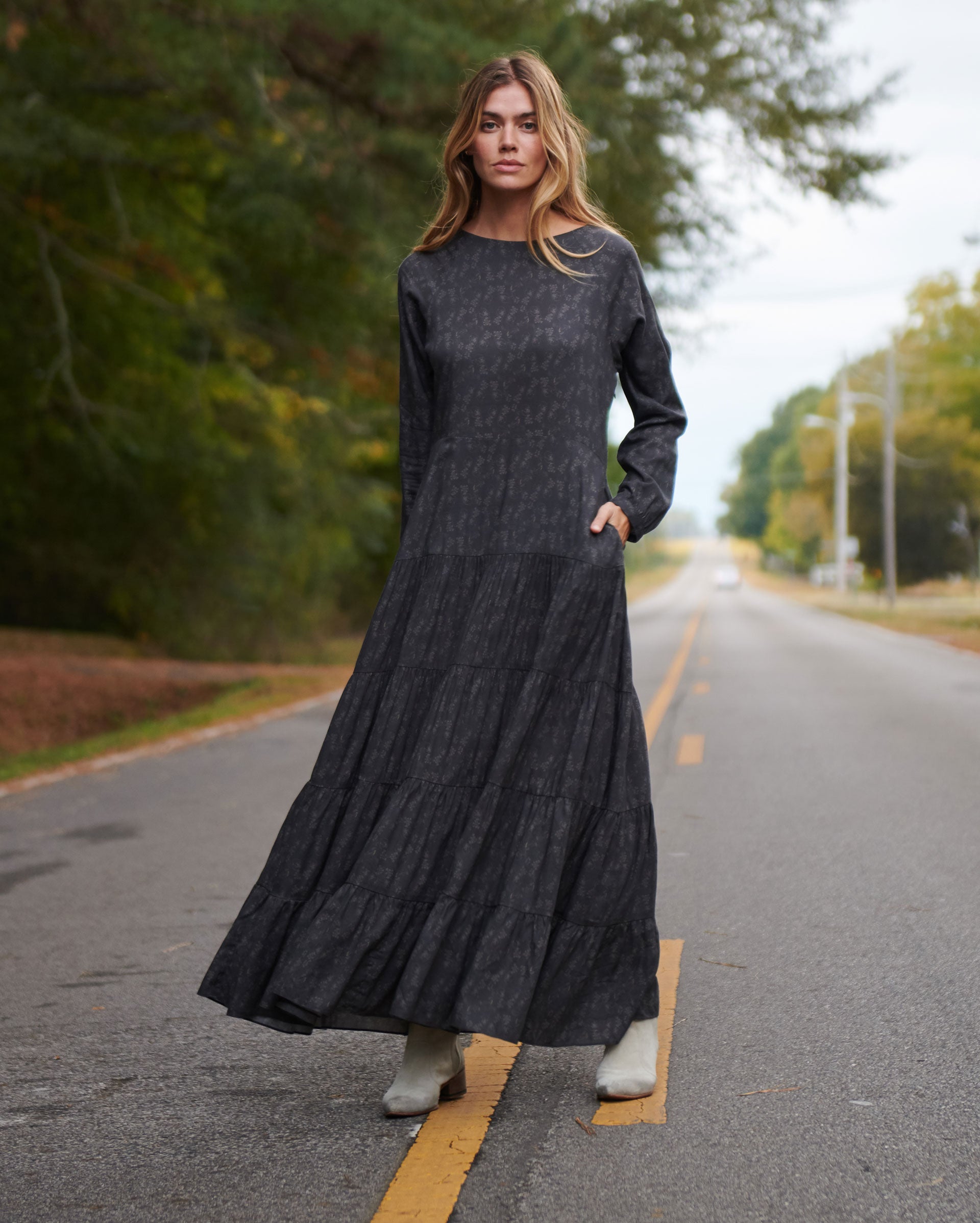 A woman in a long-sleeved, dark gray patterned maxi dress and light-colored boots stands on a road lined with trees, looking directly at the camera with one hand in her pocket.