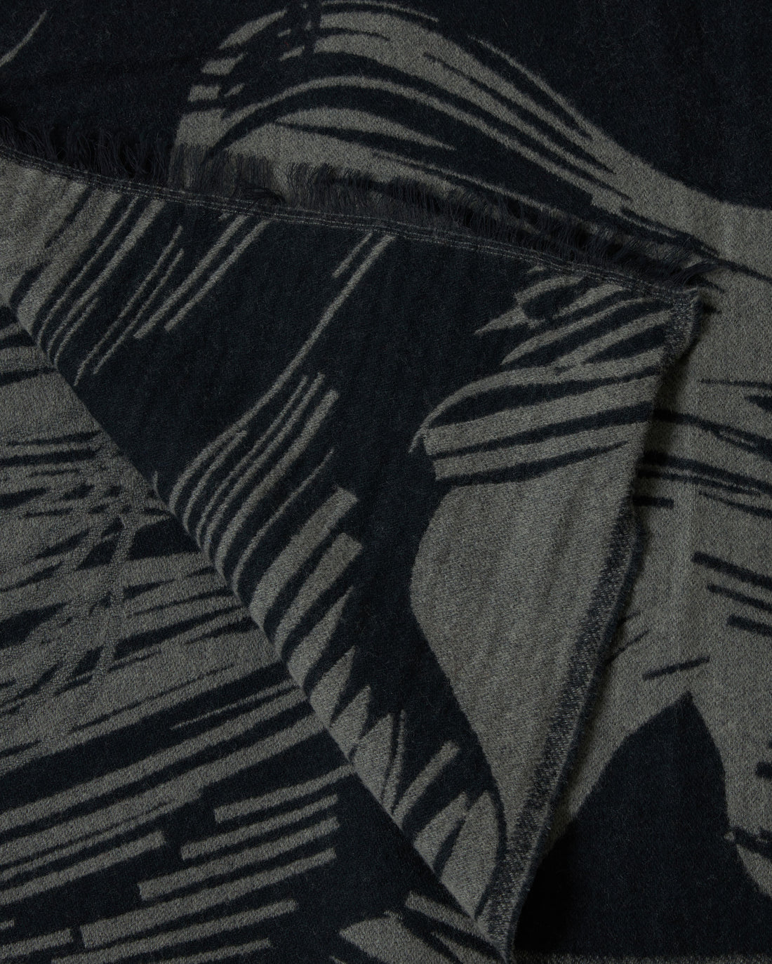 A close-up of a textured fabric with abstract black and gray patterns and fringed edges, showing a folded corner.