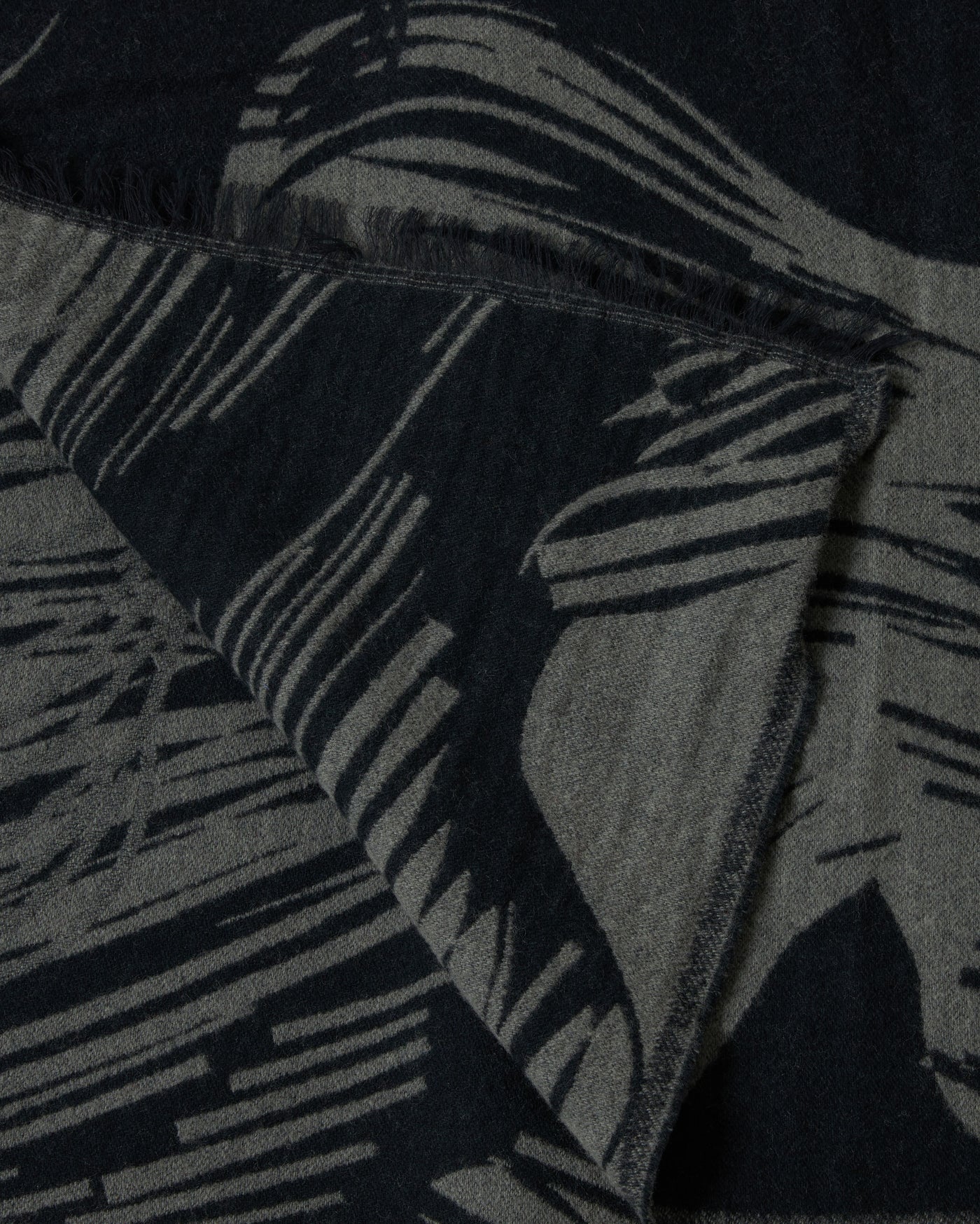 A close-up of a textured fabric with abstract black and gray patterns and fringed edges, showing a folded corner.