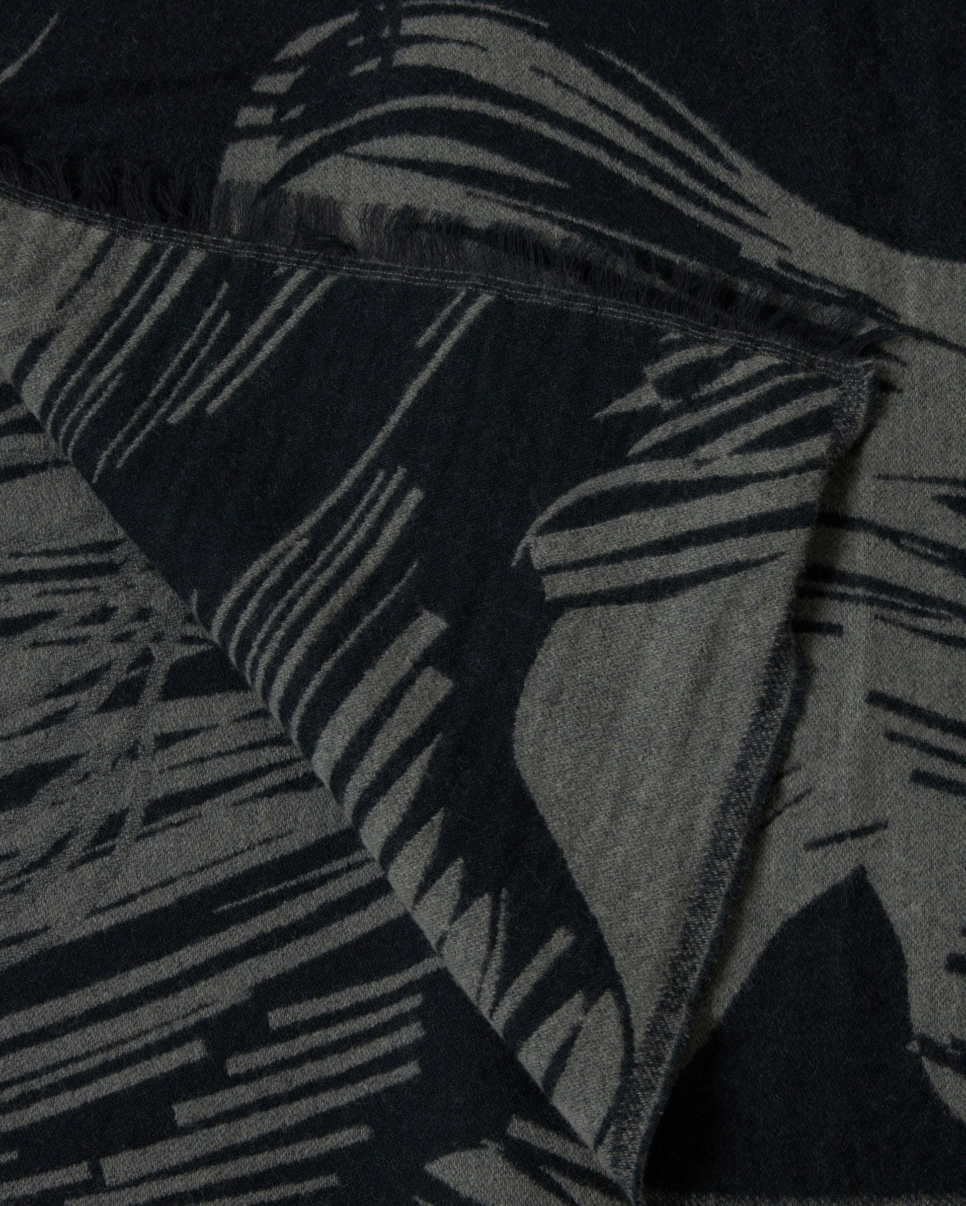 A close-up of a textured fabric with abstract black and gray patterns and fringed edges, showing a folded corner.