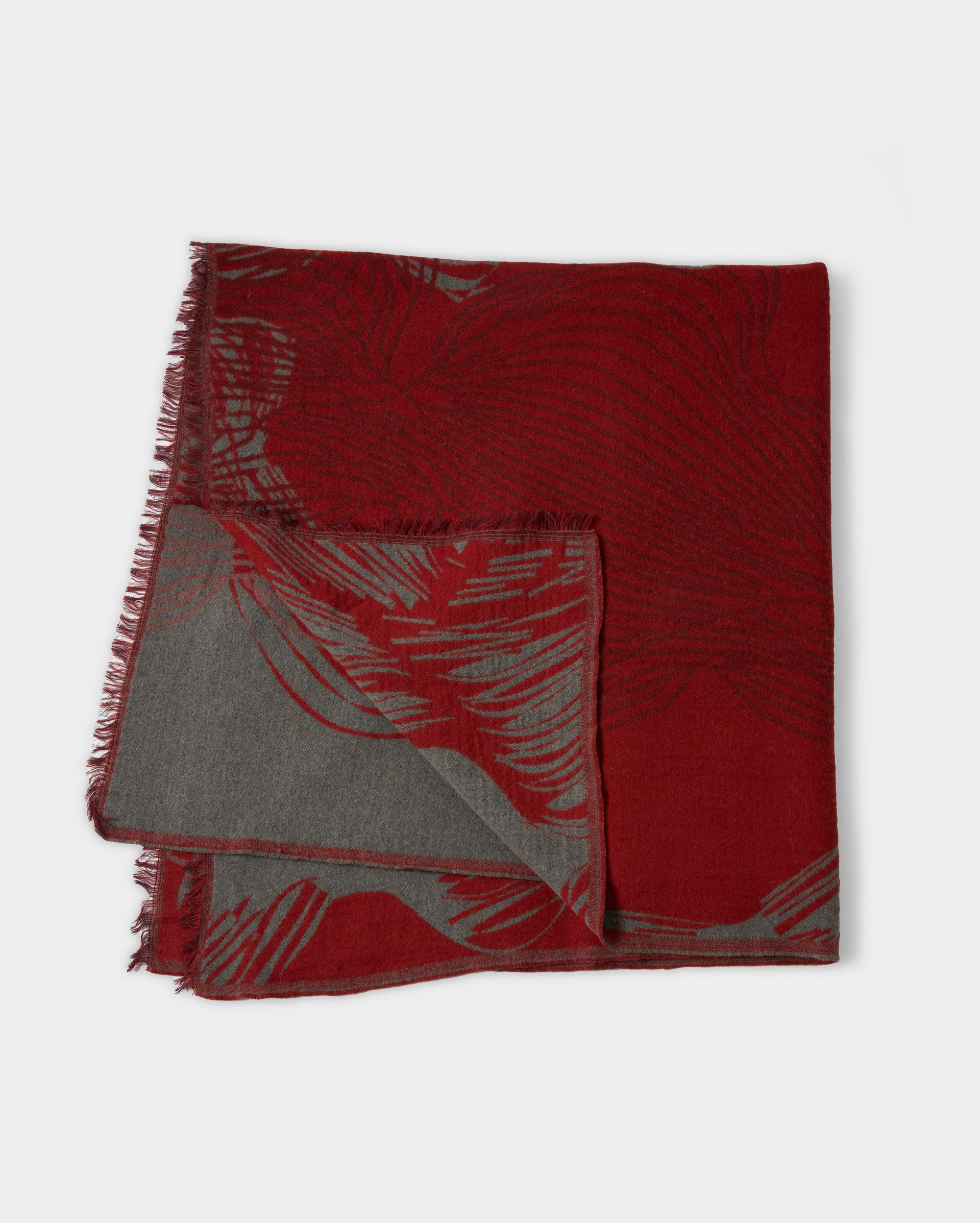A folded, rectangular scarf with fringed edges. The scarf features a red and gray abstract pattern, with the red side more prominent on top and the gray side partly visible underneath.