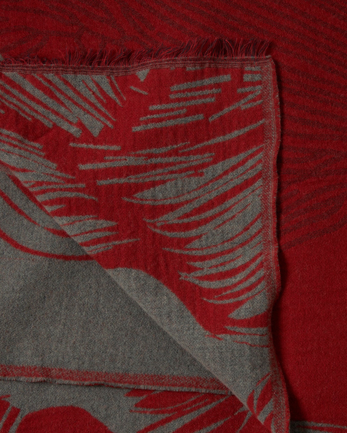 A close-up of a red and gray woven fabric with a folded corner, featuring a fringed edge and an abstract pattern resembling brushstrokes or feathers.