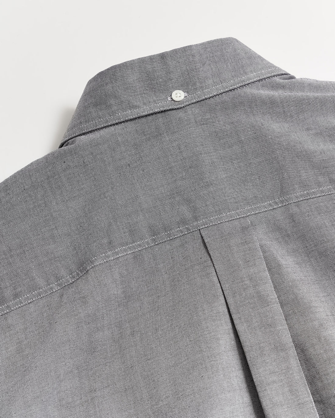 Close-up of the back of a gray button-up shirt featuring a box pleat and a small white button at the top near the collar, showcasing the shirts stitching and fabric texture.