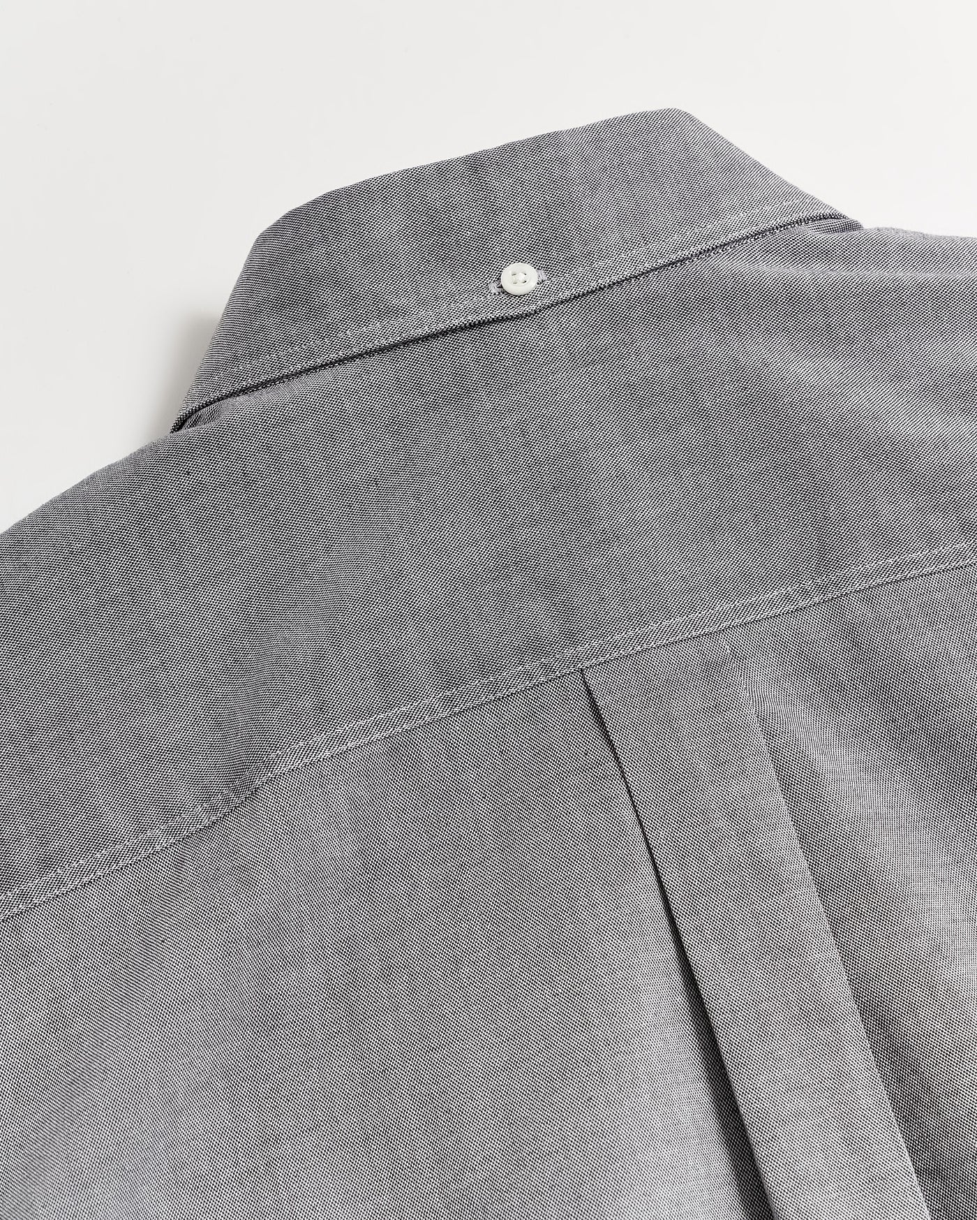 Close-up of the back of a gray button-up shirt featuring a box pleat and a small white button at the top near the collar, showcasing the shirts stitching and fabric texture.