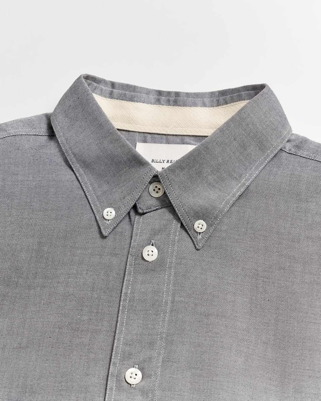 A close-up of a grey button-down shirt with a pointed collar and white buttons, displayed on a flat white surface. A label is partially visible inside the collar.