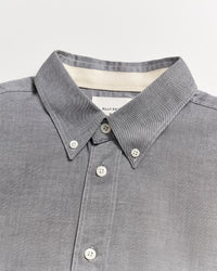 A close-up of a grey button-down shirt with a pointed collar and white buttons, displayed on a flat white surface. A label is partially visible inside the collar.