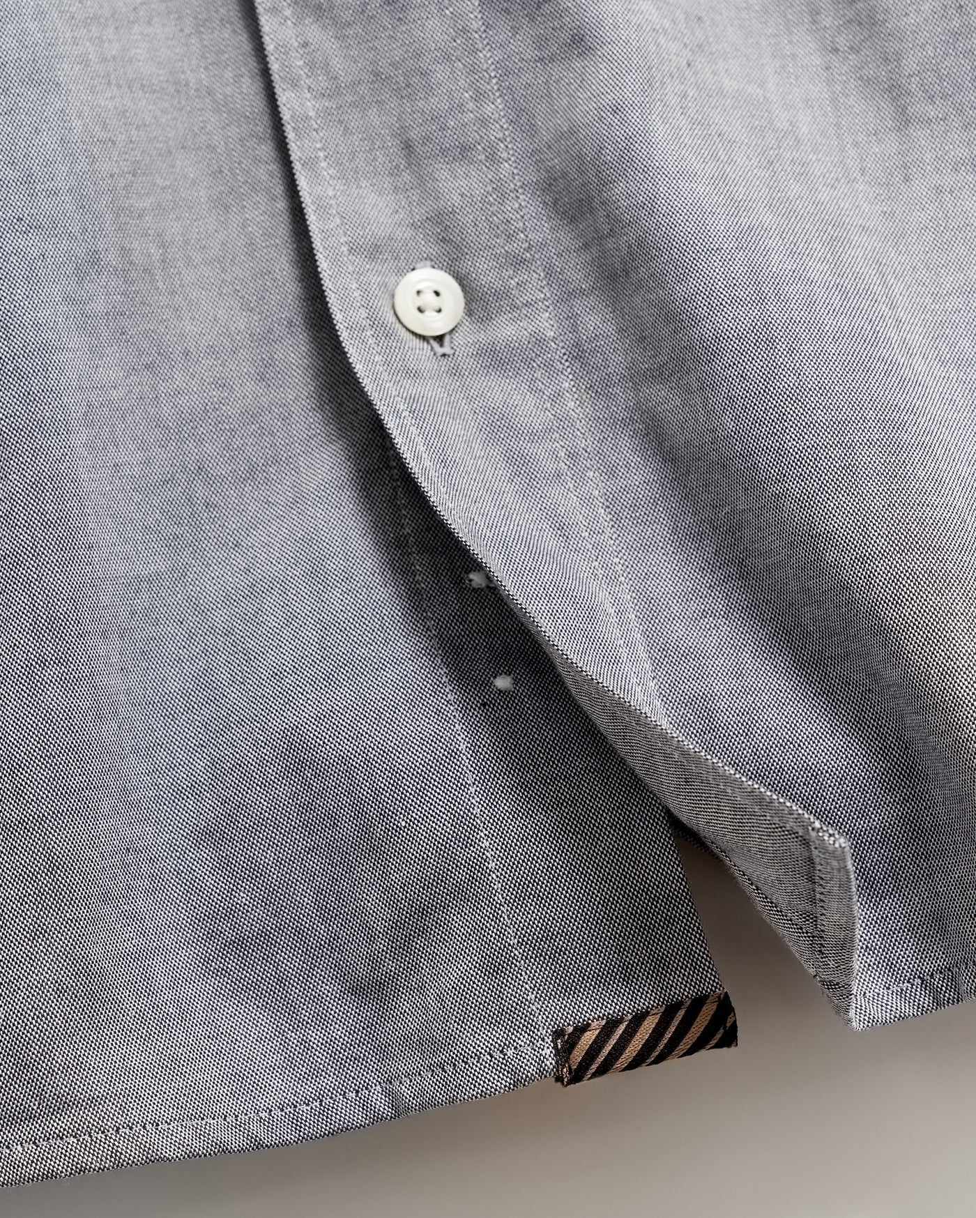 Close-up of a light gray shirt with a white button partially fastened, revealing the inside seam and a small black and brown striped fabric detail at the hem.