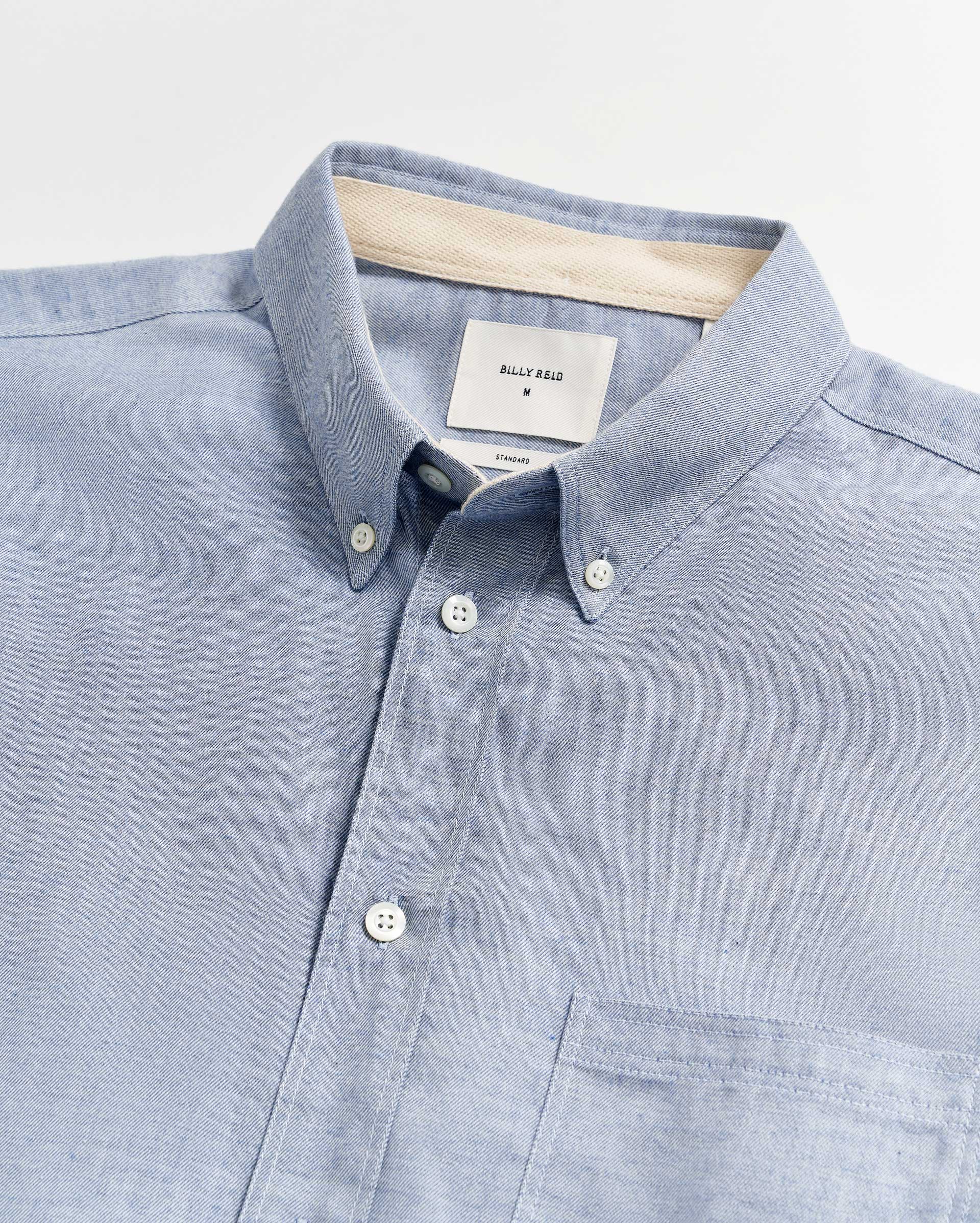 A light blue button-down shirt with a chest pocket, white buttons, and a beige inner collar strip. The shirt label inside the collar reads “BILLY REID M.”.