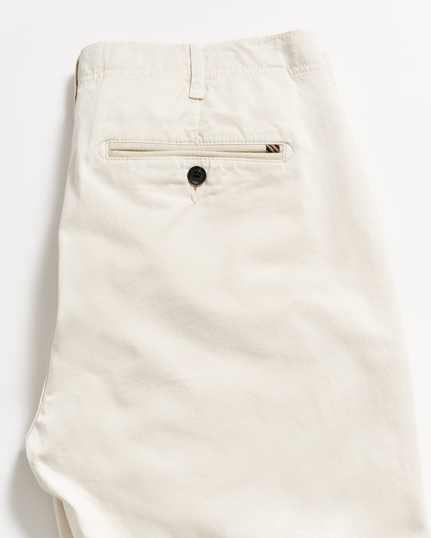 A pair of neatly folded beige pants, showing the back side with a single welt pocket featuring a black button, against a plain white background.