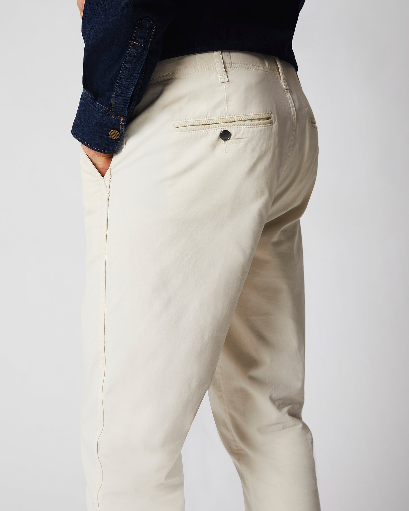 A person wearing light beige pants and a dark long-sleeve shirt with one hand in their pocket, shown from the back against a plain background.
