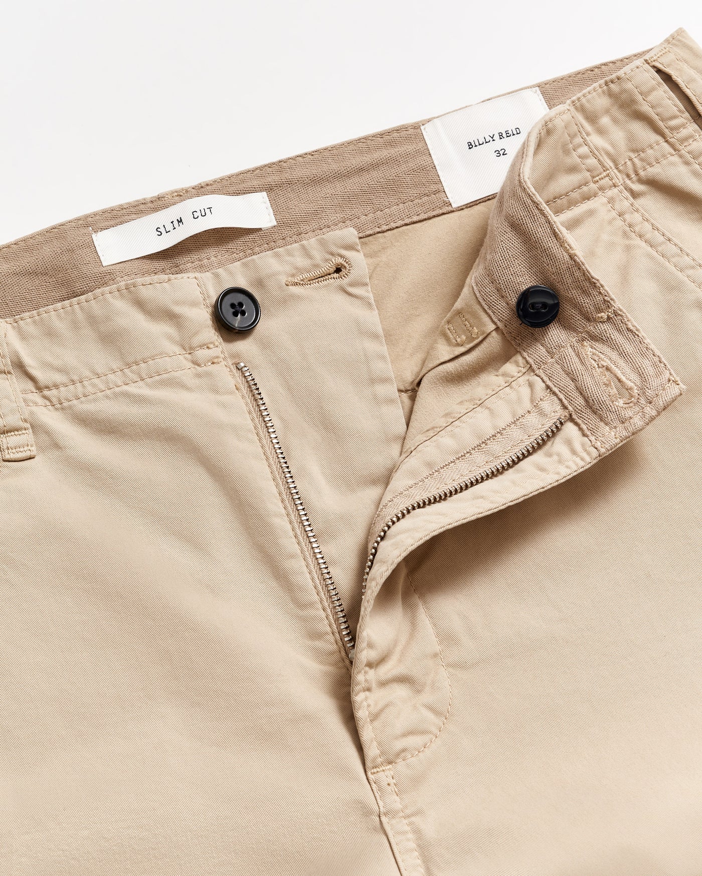 Close-up of beige slim cut trousers with the waistband partially opened, showing a button, zipper, and inner labels that read SLIM CUT and BILLY REID 32 against a white background.