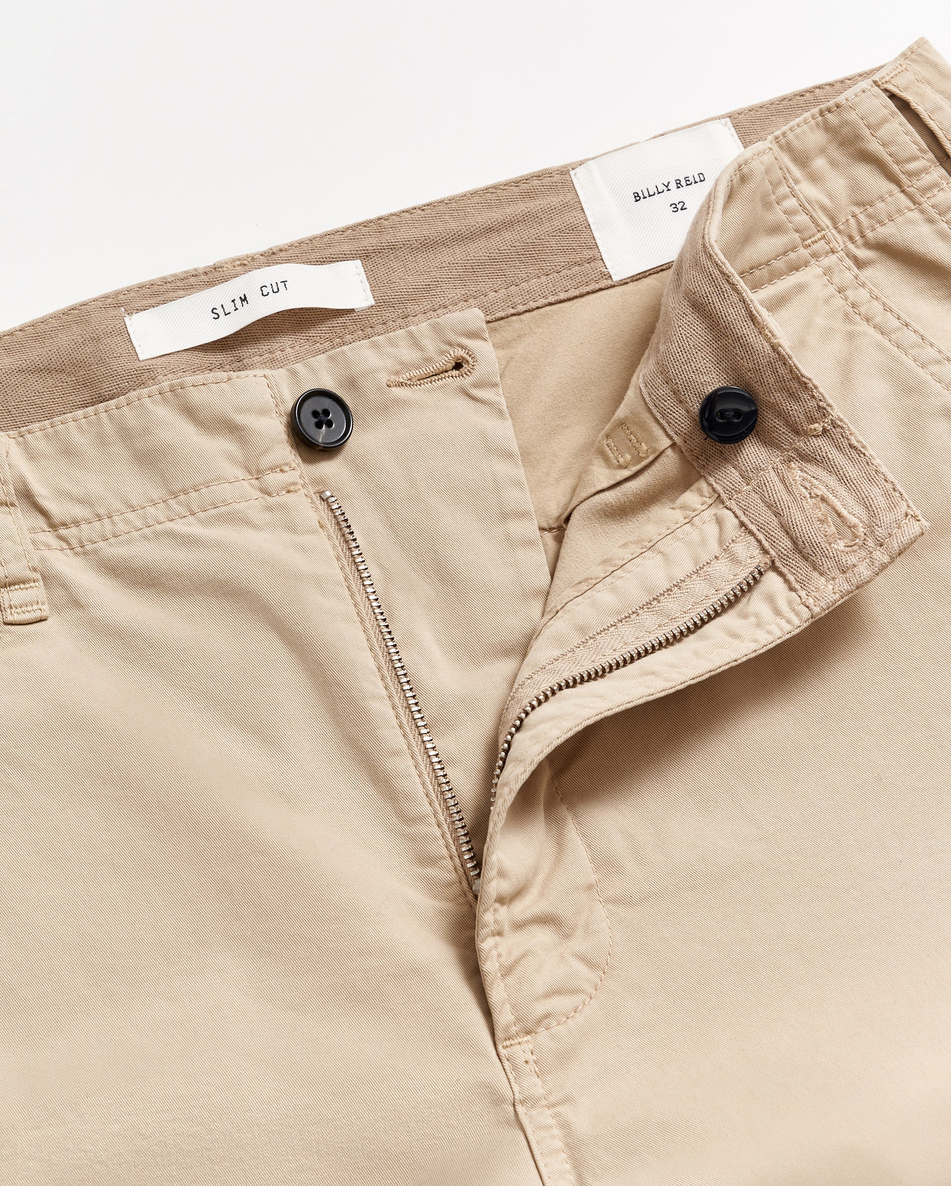Close-up of beige slim cut trousers with the waistband partially opened, showing a button, zipper, and inner labels that read SLIM CUT and BILLY REID 32 against a white background.