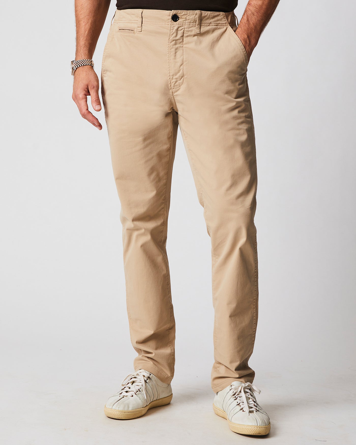 A person wearing beige chinos, a black shirt, a silver watch, and white sneakers; hands are in pockets and only the lower half of the body is visible against a plain background.